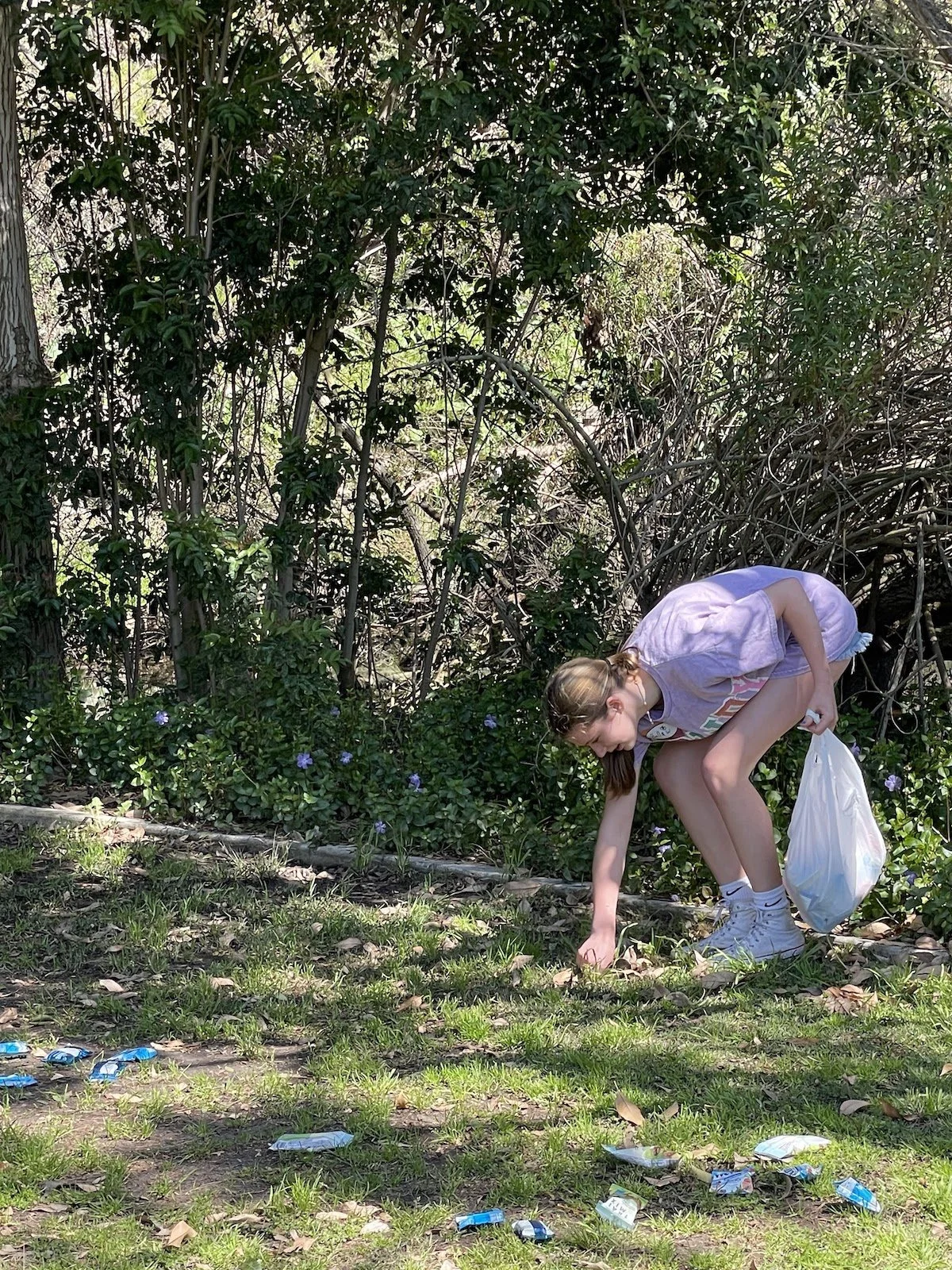A young girl crouches down to pick up litter or trash from the ground in an outdoor green setting with trees and plants and discarded trash scattered on the grass.