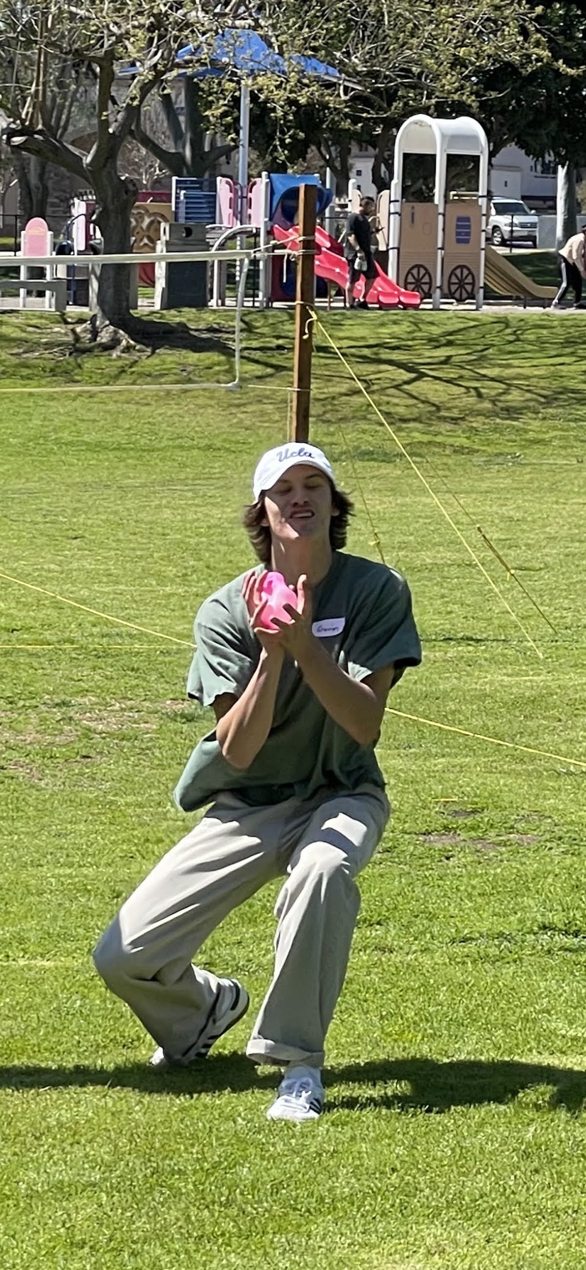 A young man wearing a UCLA baseball cap and khaki pants is kneeling on a grassy field, holding a pink object, with a playground in the background.