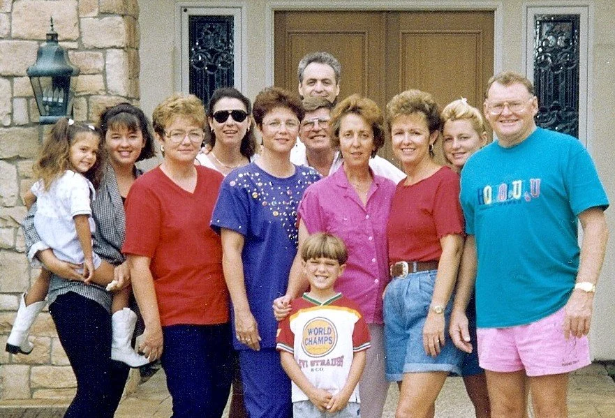 A large family standing outside a house, smiling for a group photo. The group includes children, adults, and elderly individuals, dressed casually with some women wearing brightly colored shirts and a man in a blue shirt and pink shorts.