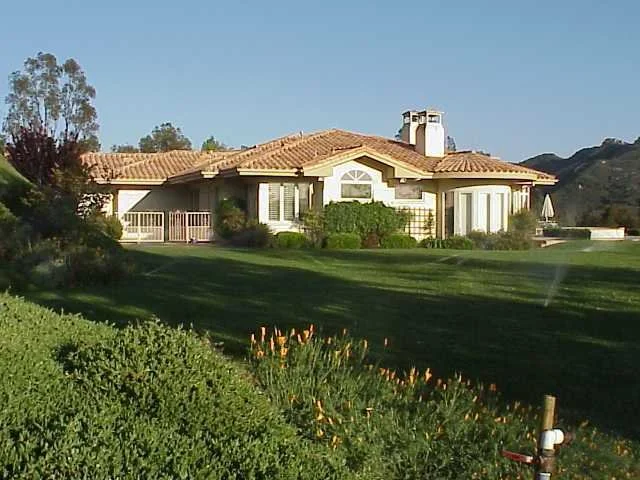 Large house with a tiled roof and a white chimney, surrounded by a green lawn and various bushes and trees, under a clear blue sky.