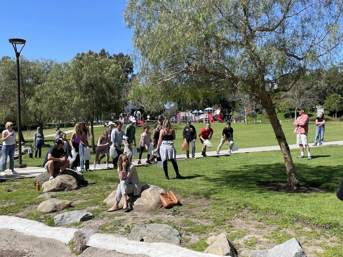 People gathered at a park on a sunny day, some sitting on rocks, others walking or standing. There are trees, a playground in the background, and a clear blue sky.