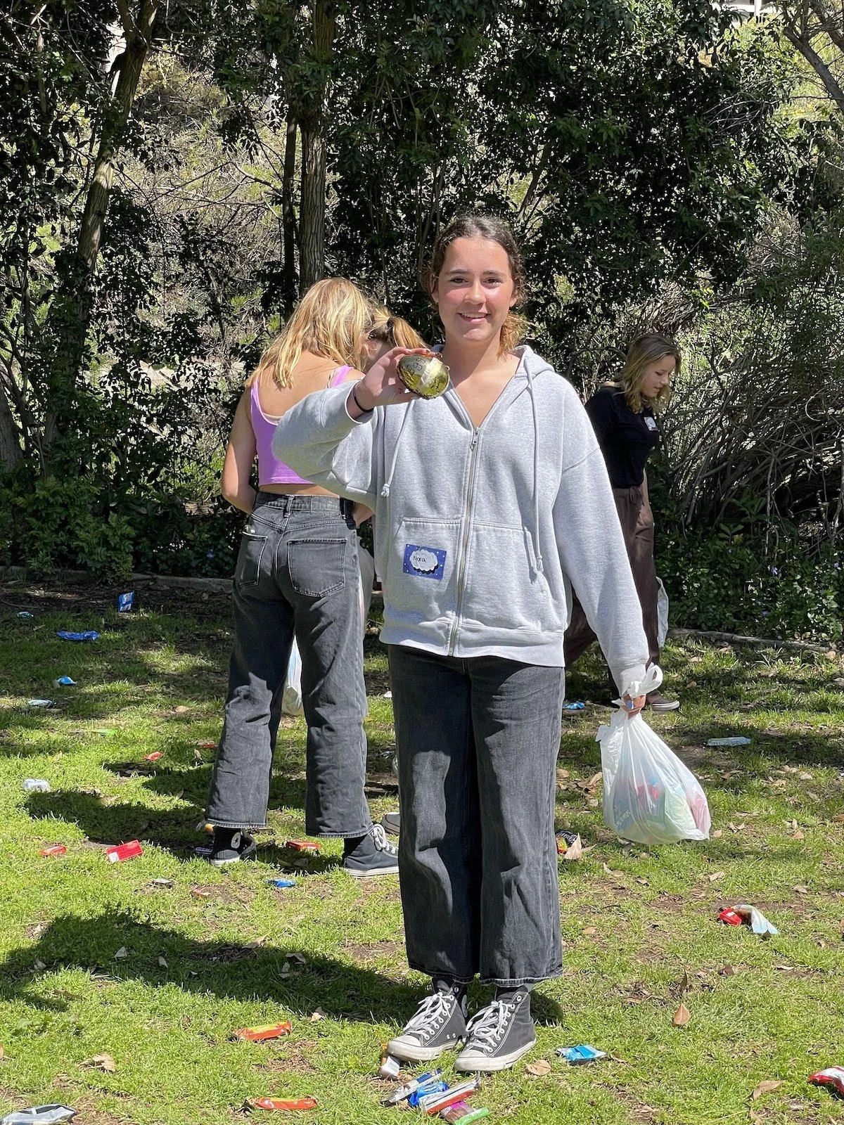 Young woman wearing a gray hoodie and gray jeans holding a trash bag and a disposable cup, standing outdoors in a wooded area with others participating in a cleanup activity, surrounded by litter on the grass.