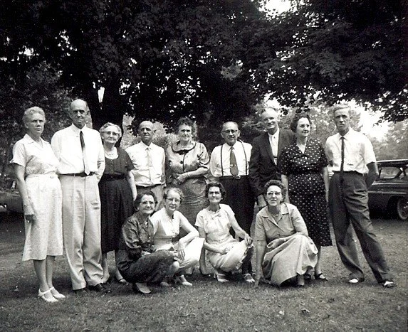 Black and white photo of fifteen people, men and women, posing outdoors in front of trees, with some cars visible in the background, during daytime.