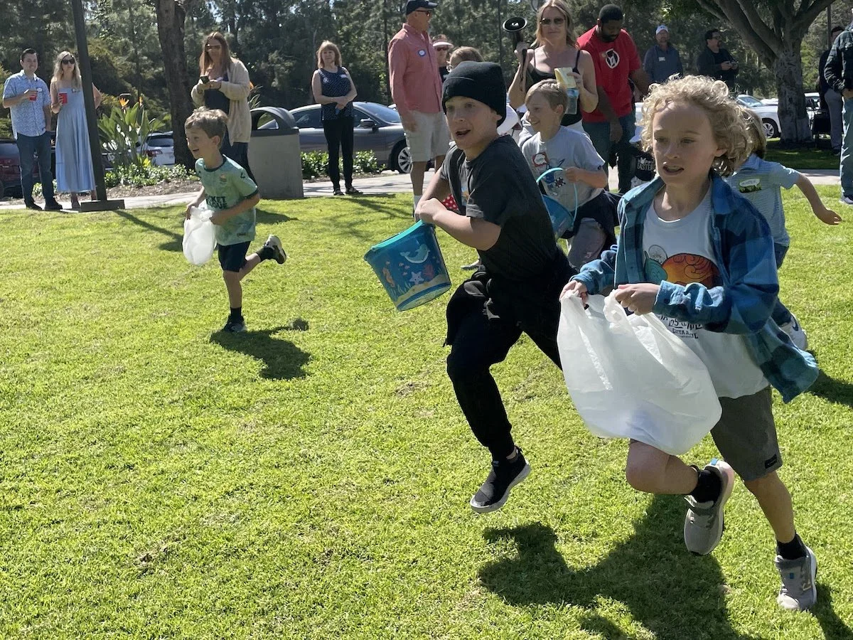 Children participating in an outdoor egg hunt, running with plastic bags and buckets to collect eggs on a grassy area, with onlookers in the background.
