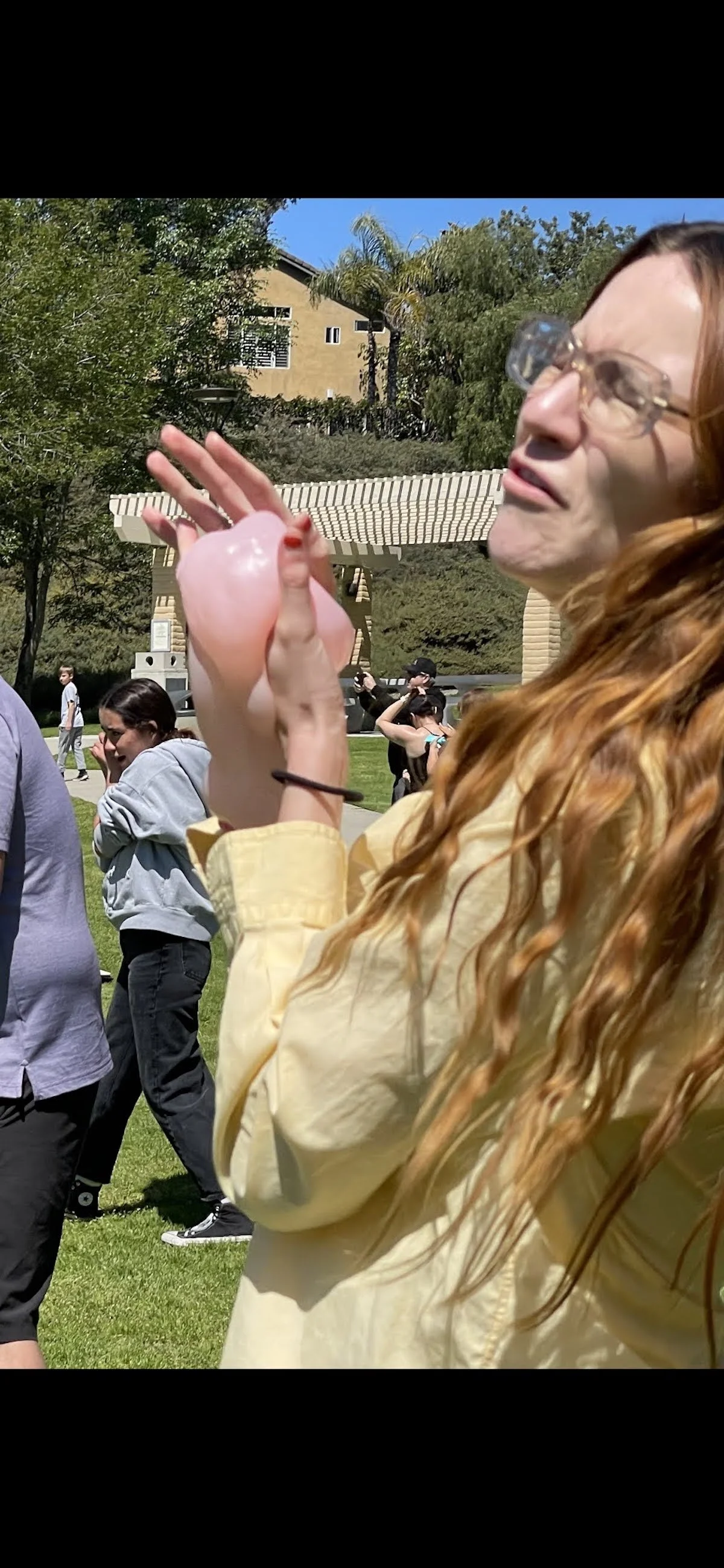 A woman with long curly hair, wearing glasses and a yellow jacket, holding a pink cotton candy at an outdoor event on a sunny day.
