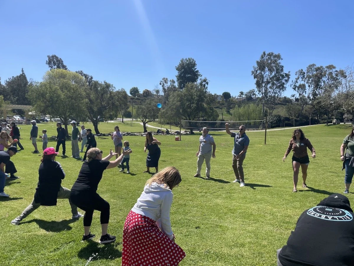People outdoors in a park, some squatting or standing, participating in a social gathering or activity on a sunny day with trees and blue sky in the background.