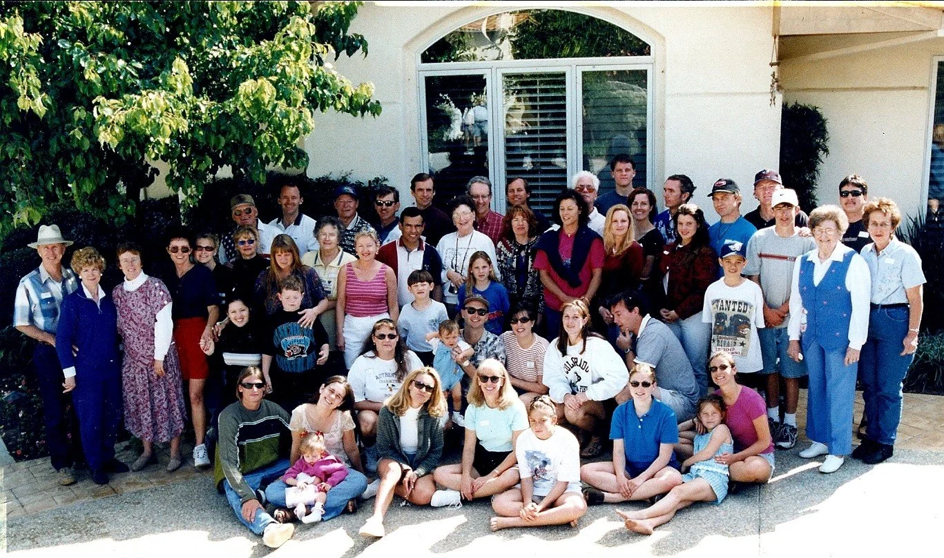 Large group of people of various ages posing outdoors in front of a house with large window and greenery, likely at a social gathering or family event.