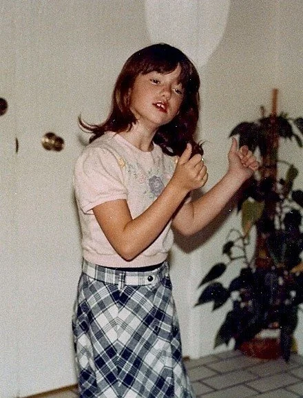 Young girl with shoulder-length brown hair, wearing a light pink t-shirt and plaid skirt, standing indoors near potted plants.