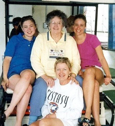 Four women sitting together indoors, smiling, with large windows behind them. The woman in the center is older, wearing a yellow jacket, and has gray curly hair. The three younger women surrounding her are wearing casual clothing, with two of them in