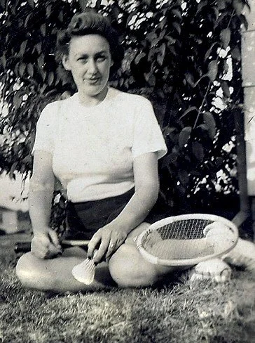 A woman sitting cross-legged on the grass with a tennis racket resting on her lap, smiling at the camera, with leafy bushes in the background.