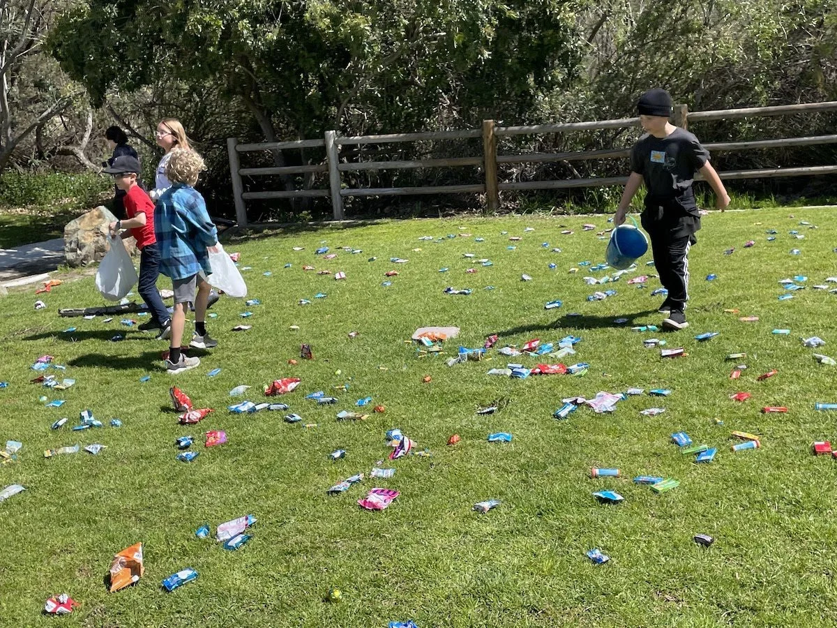 Children participating in a candy hunt outdoors on a grassy field, surrounded by trees and a wooden fence.