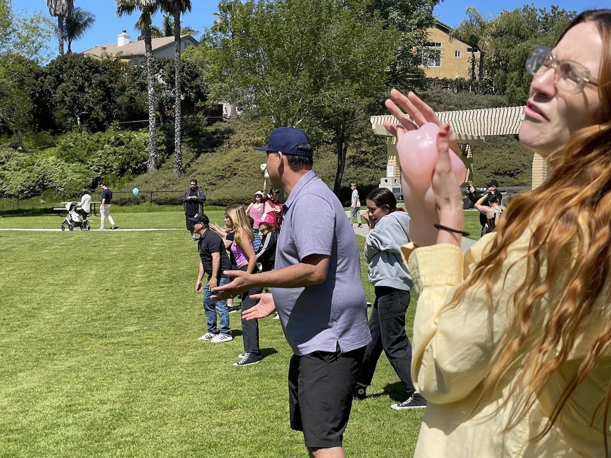 People standing on a grassy field with trees and houses in the background, some talking or using phones, and a woman in the foreground holding a pink balloon.