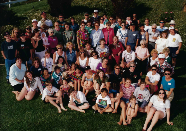 A large group of people, including children, adults, and seniors, gathered on a grassy area outdoors for a group photo on a sunny day.