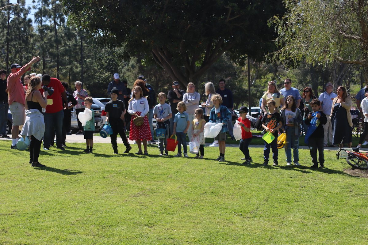 Children and adults standing on a grassy field outdoors, possibly participating in a group activity or game, with trees and parked cars in the background.