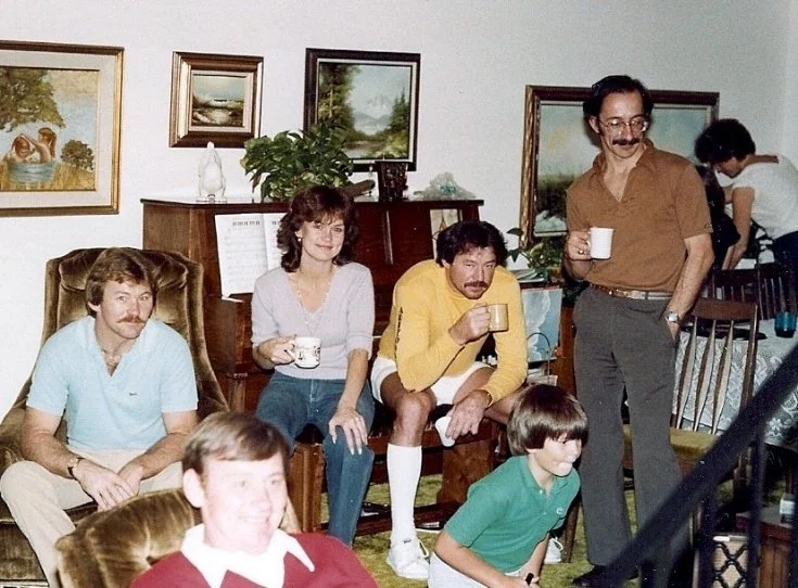 Five people sitting and standing in a living room, holding mugs, with paintings on the wall and a piano in the background.