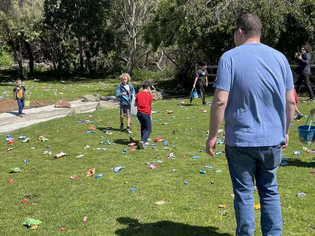 Children and adults participating in an egg hunt on a grassy field with trees in the background, scattered with colorful plastic eggs.