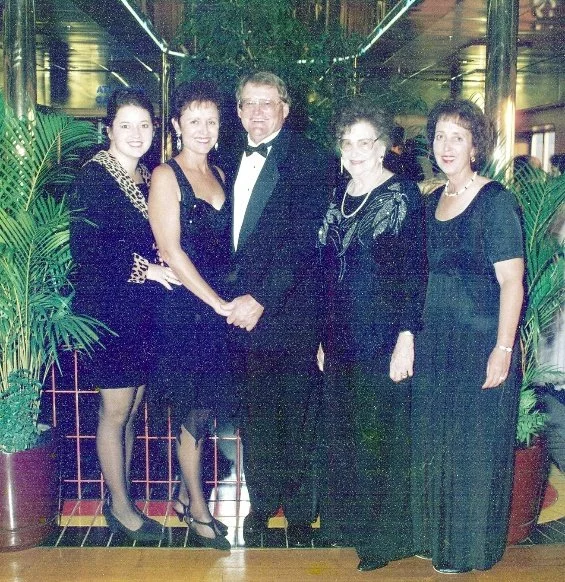 Five people dressed in formal attire smiling in front of lush green plants at a social event in a decorated venue.
