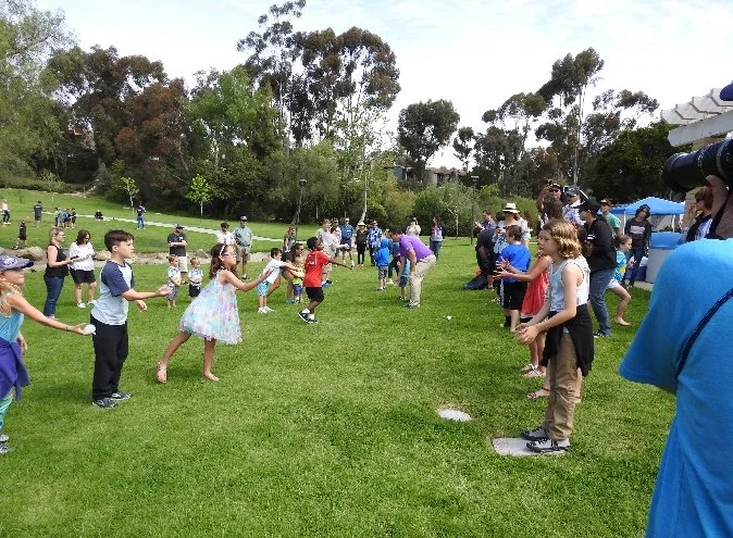 Children and adults participating in a tug-of-war game on a grassy field in a park, with trees and spectators in the background.
