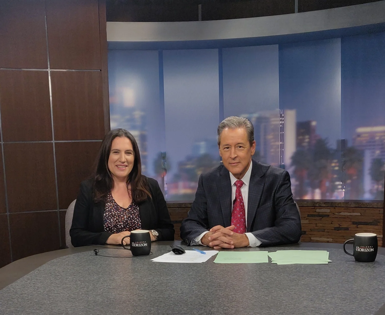 Two news anchors, a woman and a man, sitting at a news desk with papers and coffee mugs that say 'Arizona Horizon,' in a television studio with a cityscape backdrop.