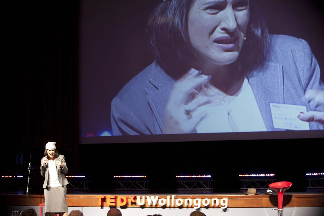 A woman in a business suit and nurse's cap giving a TED Talk on stage, with a large screen behind her displaying a close-up of her expression suggesting she is emotionally distressed.