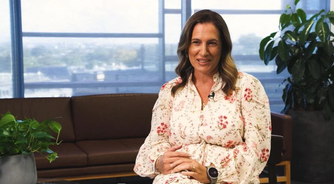A woman sitting indoors in front of large windows with city view, wearing a floral blouse and a smartwatch.