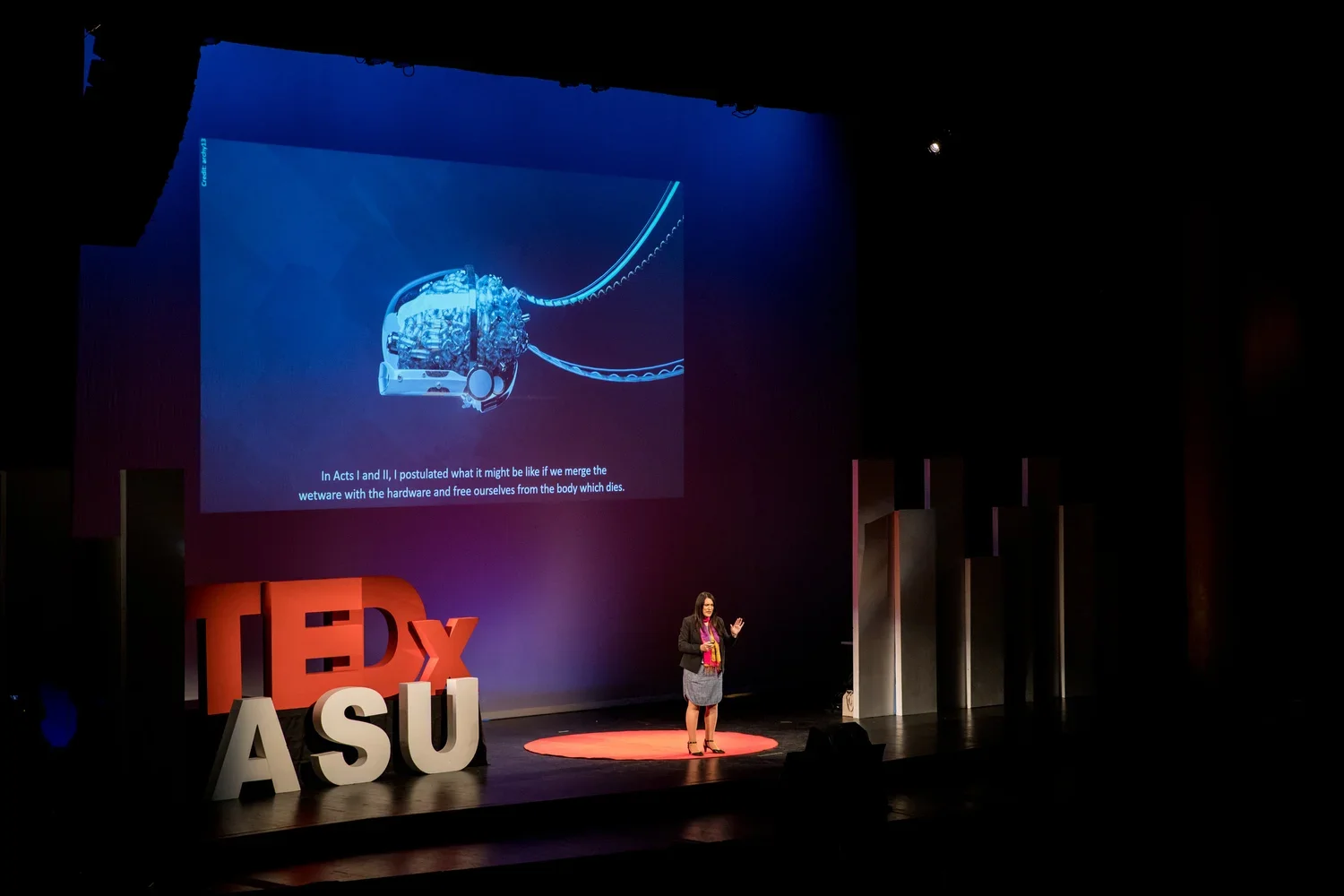 A woman is giving a presentation on a stage at TEDxASU, with a large screen displaying a scientific diagram of a mechanical device and some text. The stage has large red and white letters spelling 'TEDx ASU' and  is mostly dark with spotlights on the speaker.