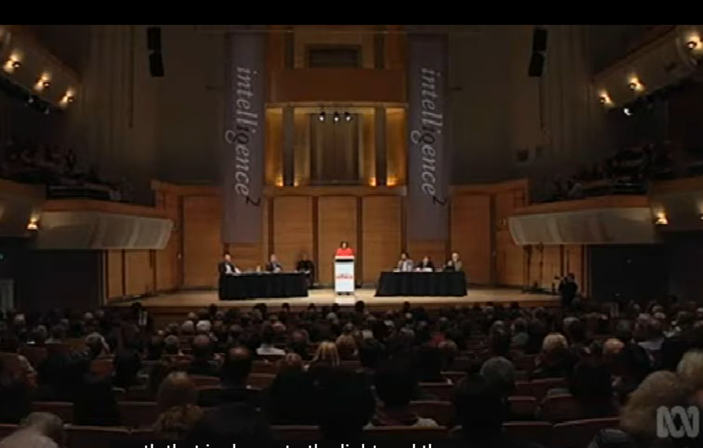 A panel of people seated at tables on a stage in an auditorium, with a speaker at a podium in the center. The auditorium is filled with an audience. Two large banners hang behind the stage with the words 'little
gesturez' printed on them.