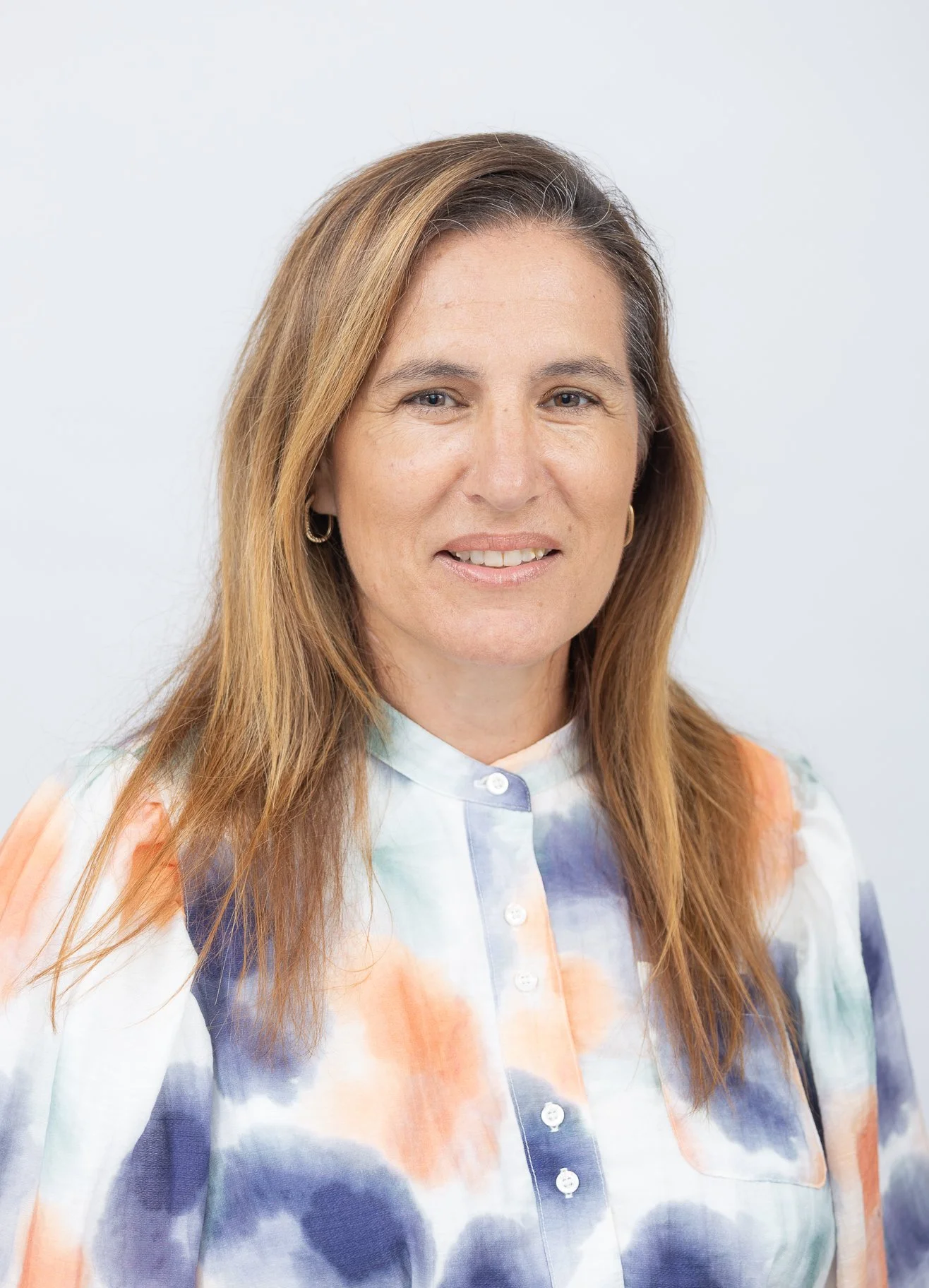 A woman with long light brown hair and hoop earrings smiling against a plain white background, wearing a colorful tie-dye button-up shirt.