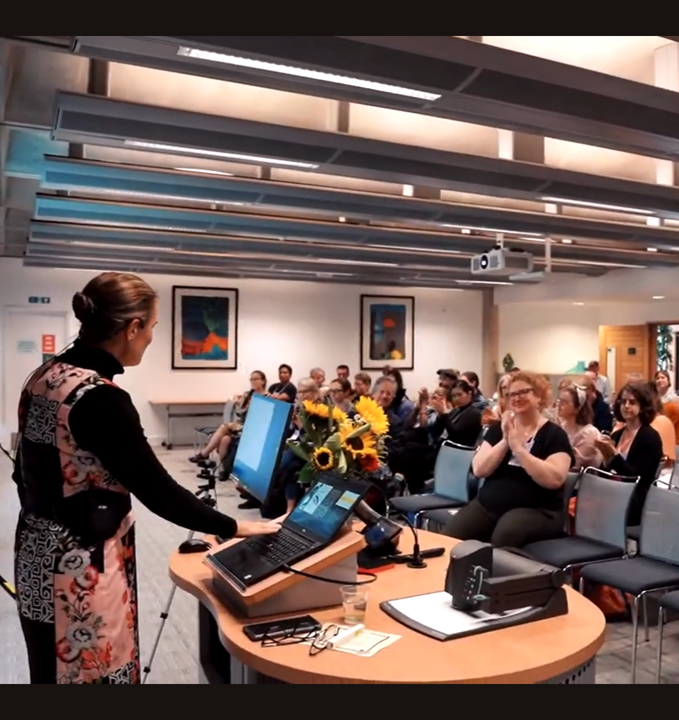 Katina giving a keynote presentation to an audience seated in a conference room at Cambridge University, with the audience reacting positively. The room has modern decor, ceiling-mounted lighting, and framed artwork on the walls.