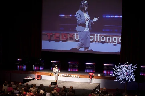 Woman in business attire giving a presentation on stage at TEDx Wallongong, with a large screen behind her displaying her image.