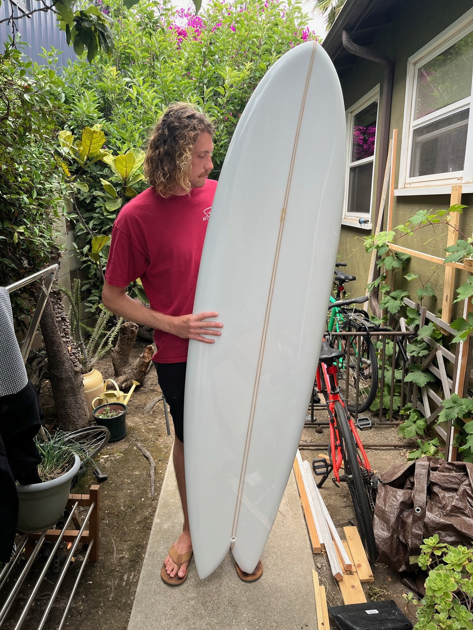 A man with curly blond hair and a red shirt is standing outdoors in a garden, holding a white surfboard upright. He is wearing sandals, and there are plants, bicycles, and gardening tools around him.