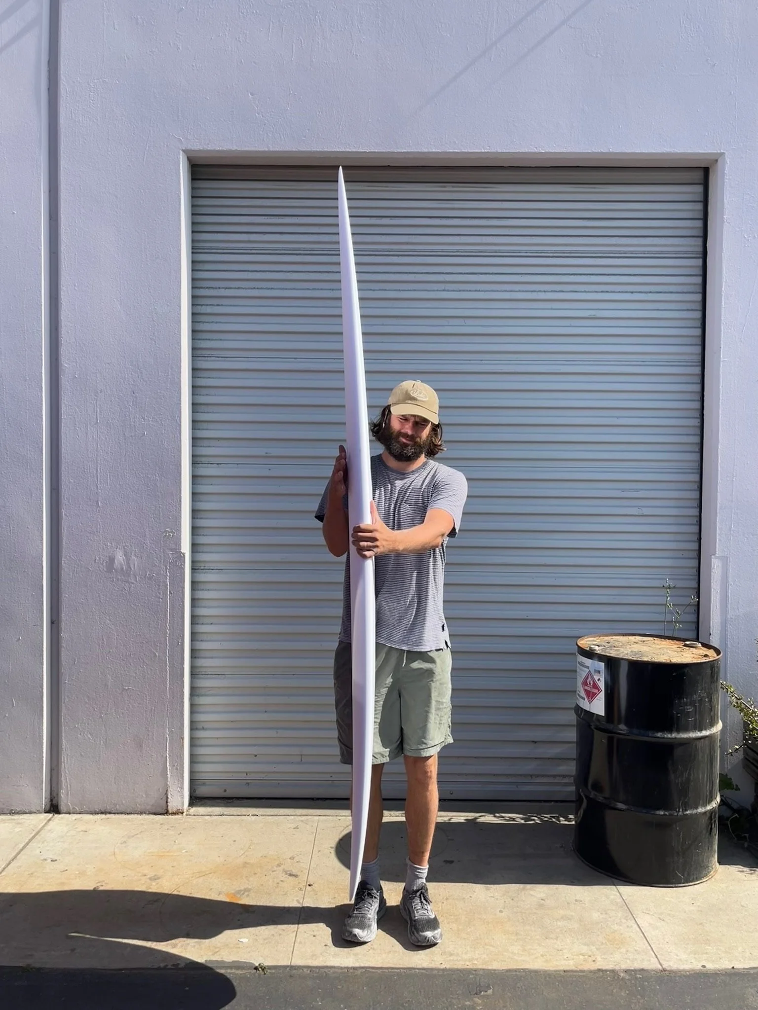 A man with long hair and a beard, wearing a beige cap, gray striped t-shirt, and shorts, is holding a long white tube vertically in front of him. He stands on a concrete sidewalk in front of a closed gray metal roll-up door, with a black trash barrel nearby and a white wall to the left.
