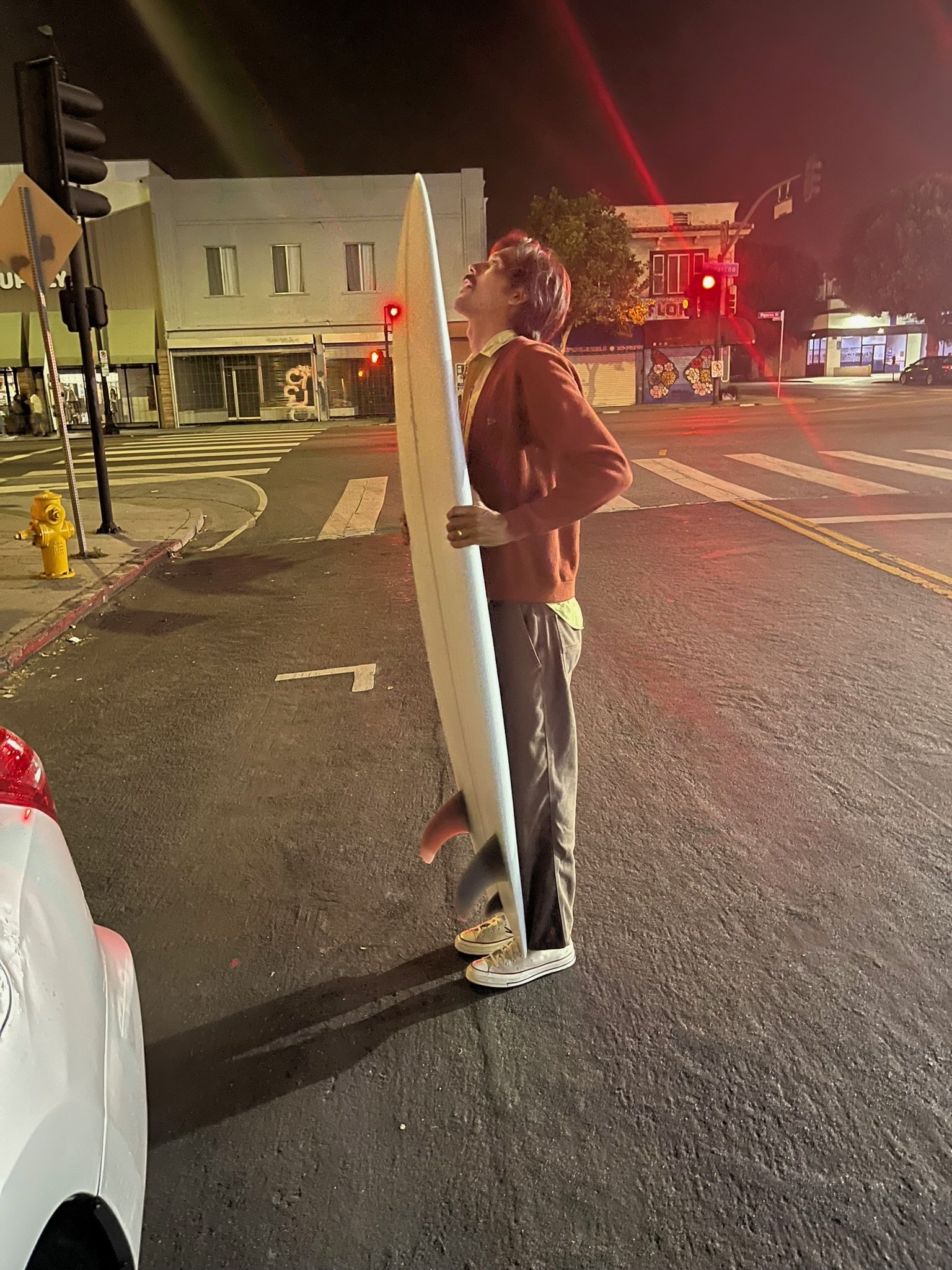A person holding a surfboard on a city street at night. They are wearing a brown jacket, beige pants, and white sneakers. The street is illuminated by traffic lights and other streetlights, with some shopfronts visible in the background.