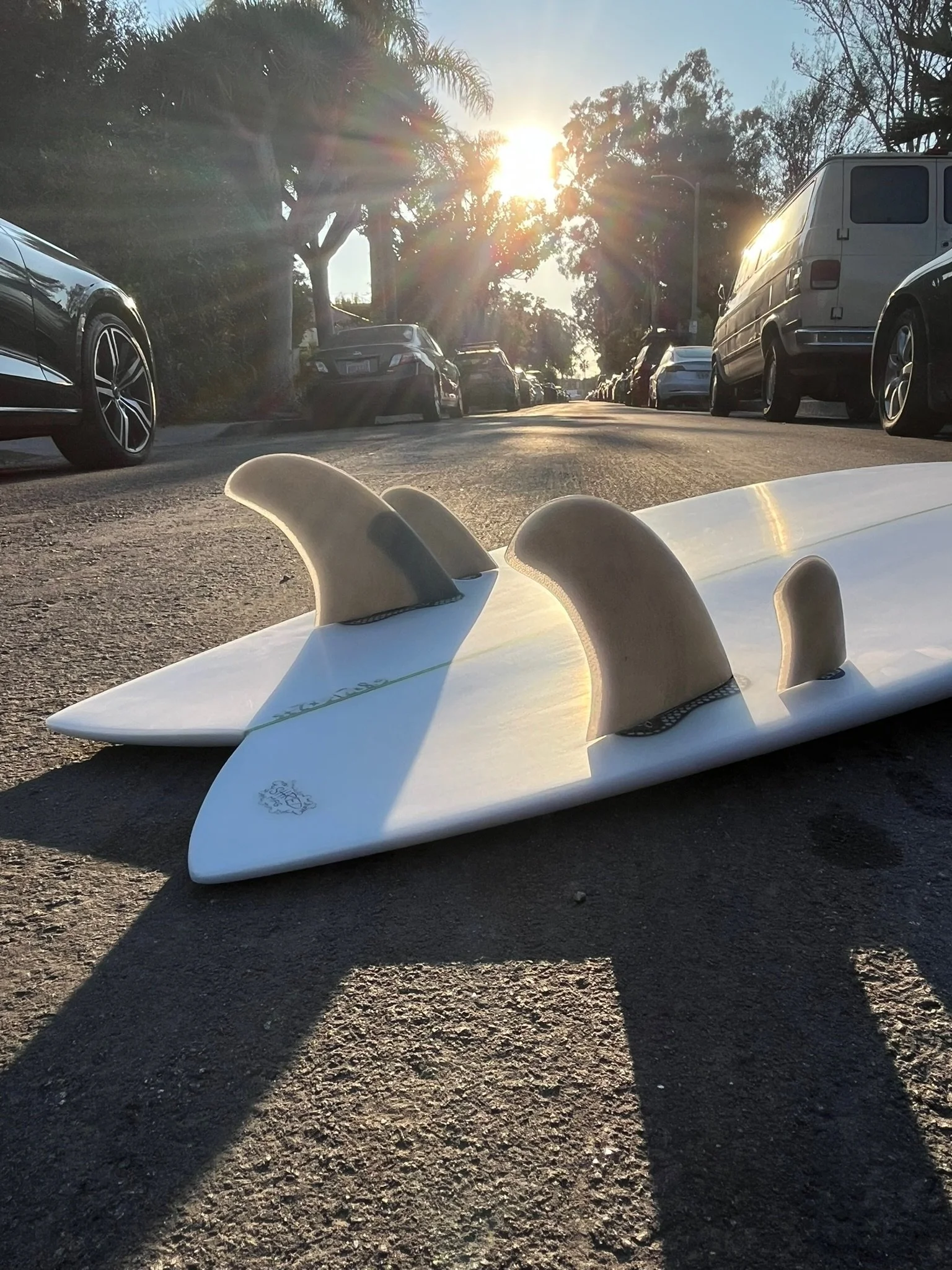 A white Quad style surfboard with fins lying on the ground in a parking lot, with parked cars on either side, trees, and the sun setting in the background.