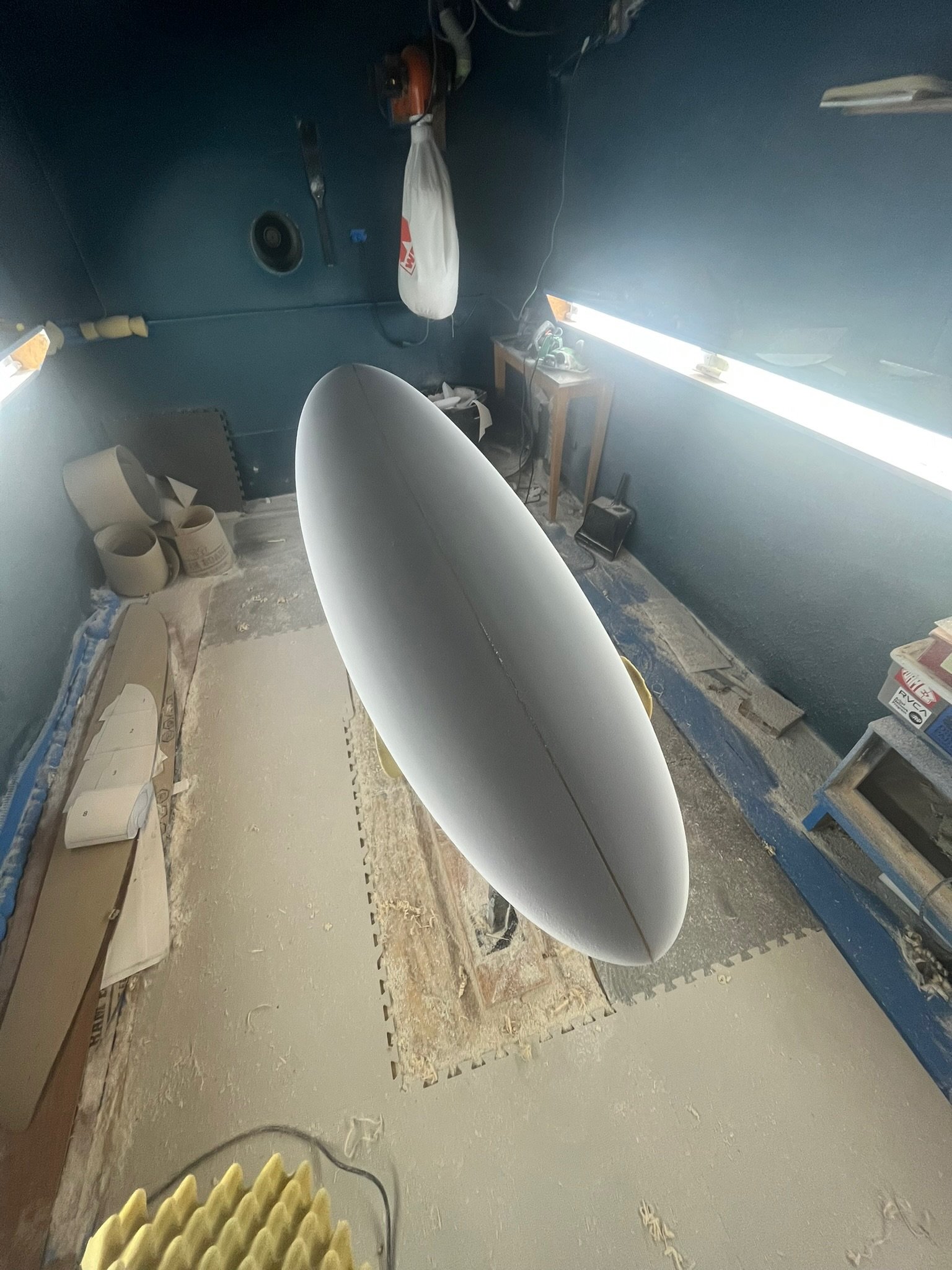 A surfboard in a surfboard shaping room with various tools, a heater, and a work table, indicating a space dedicated to surfboard crafting.
