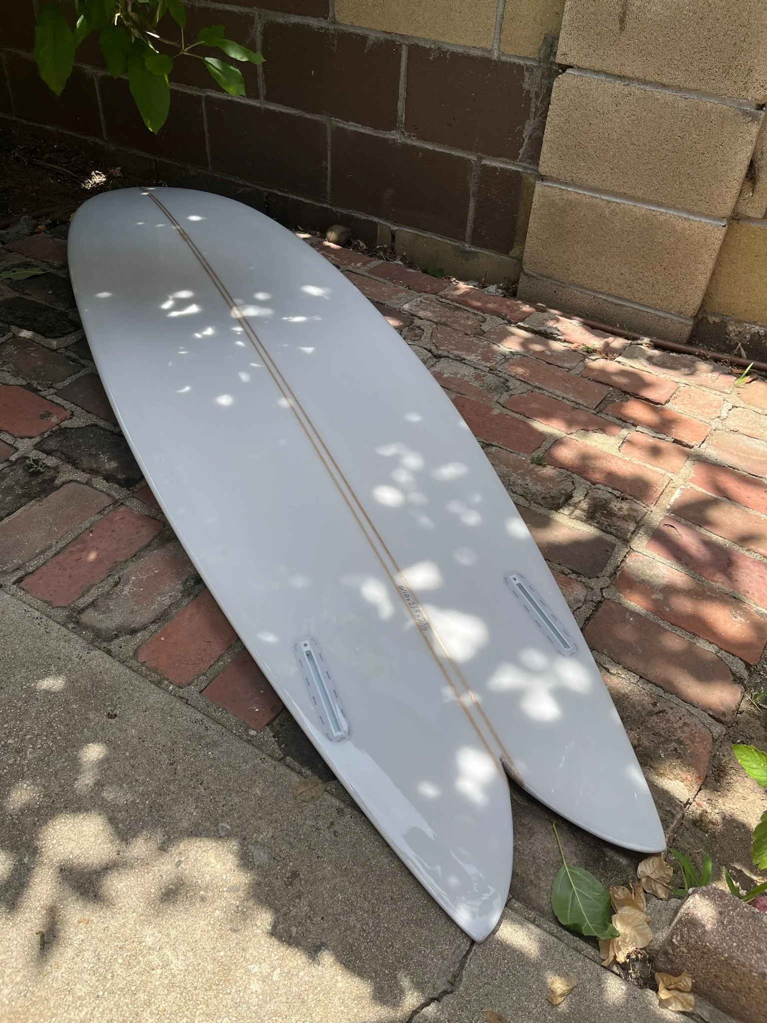 Swallow tail surfboards resting on a brick pathway, partially shaded by leaves, against a brick wall.
