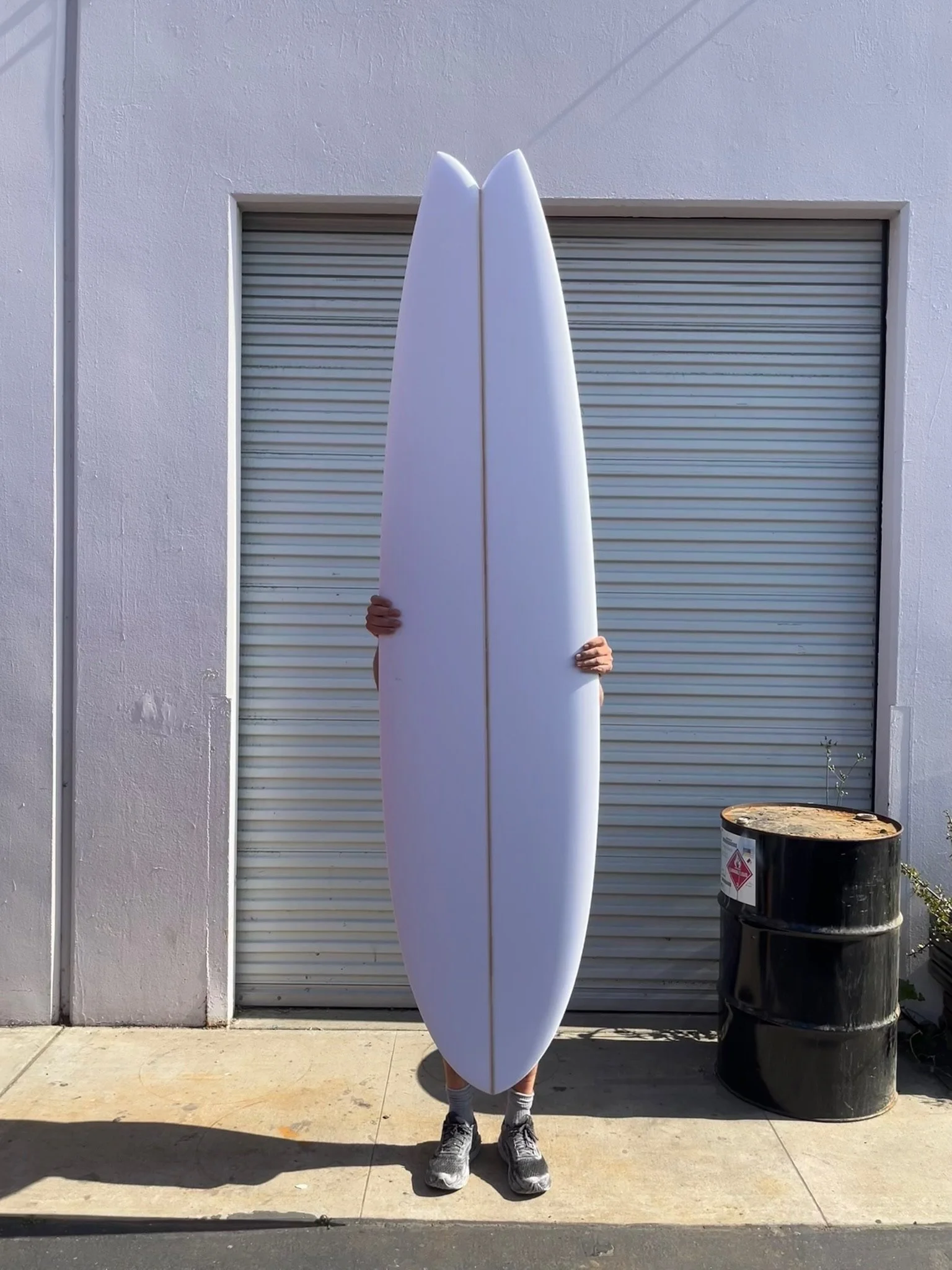 Person holding a white long fish surfboard standing in front of a garage door with a black barrel nearby.