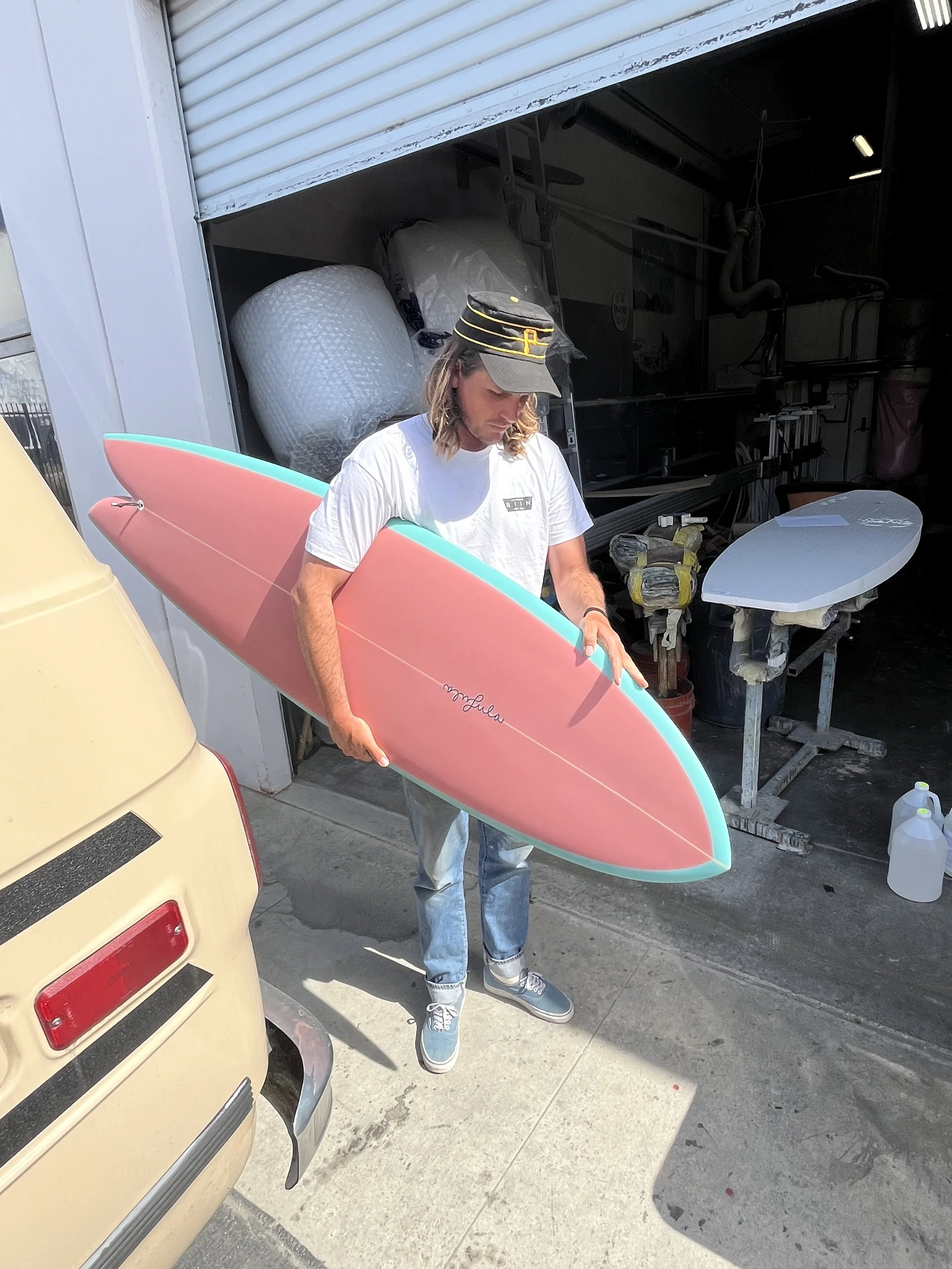 A man with long hair wearing a hat and white t-shirt holding a pink and turquoise surfboard. He stands outside near a yellow van, with a garage door open behind him showing shelves, tools, and surfboard equipment.