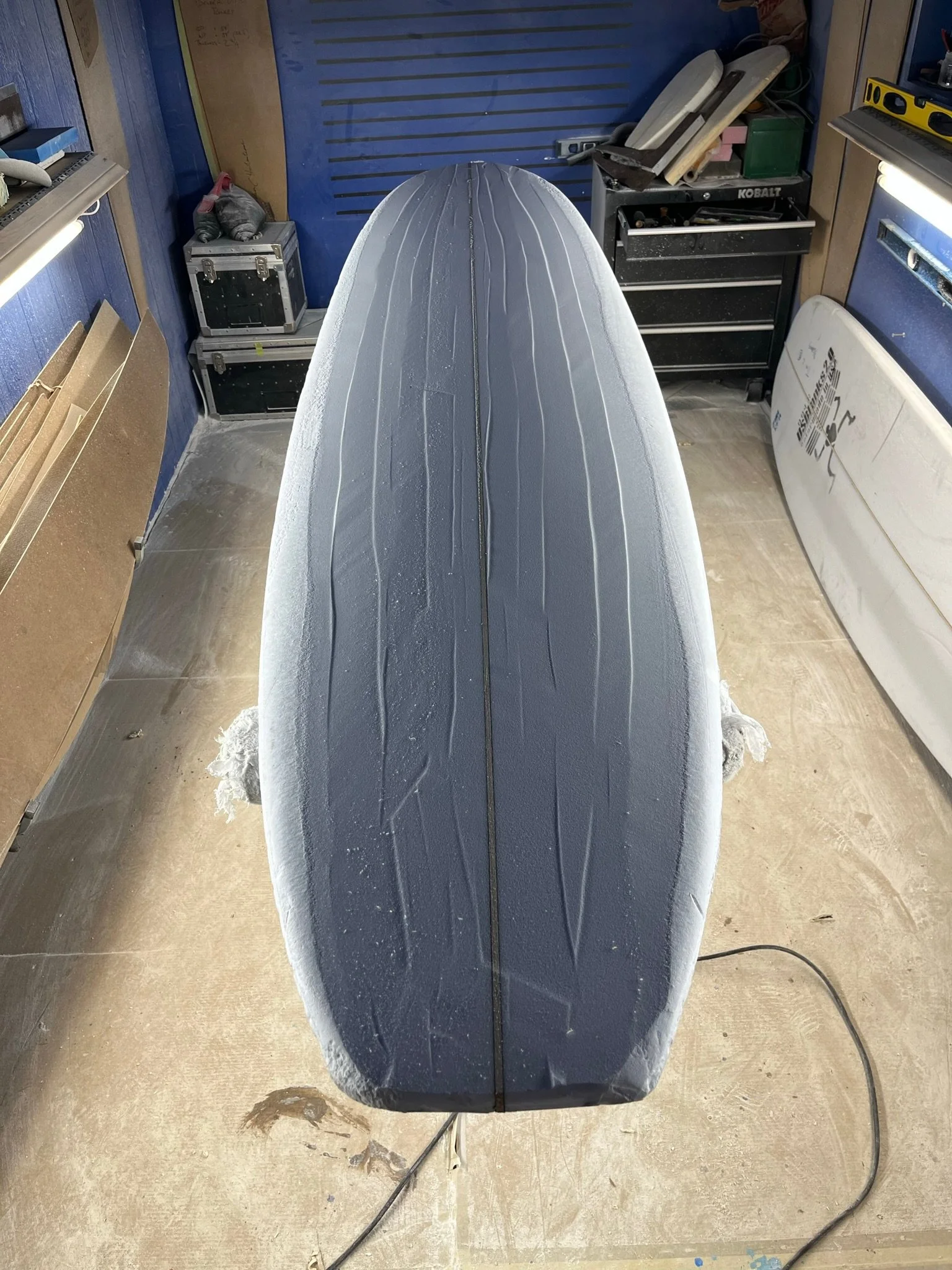 A surfboard with a fin on the bottom, positioned vertically in a workshop or garage, with various tools and equipment around.
