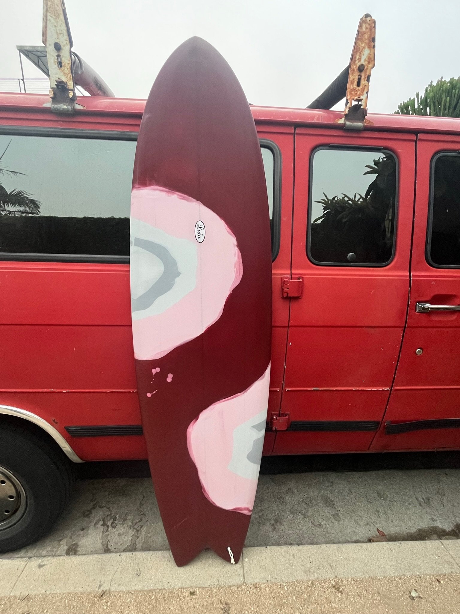 A red vehicle with a rusty roof rack and a large pink and brown surfboard leaning against the side, with the surfboard partially concealing the windows and door of the vehicle. The surfboard has a small white sticker with the name 'Lulu' on it.
