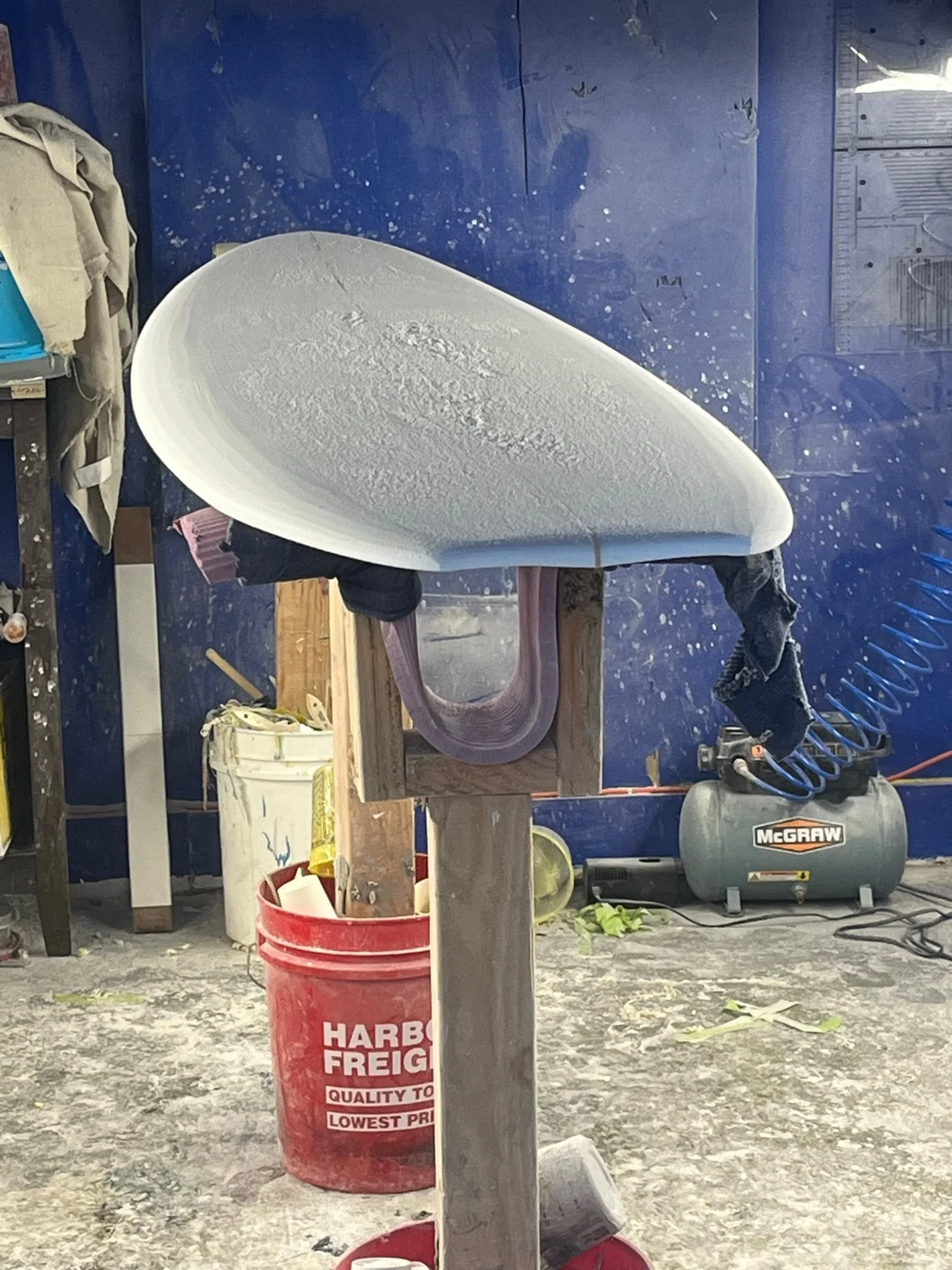 A workshop with a large surfboard-shaped object being built or repaired, surrounded by tools and supplies, against a blue wall with splatters of paint.