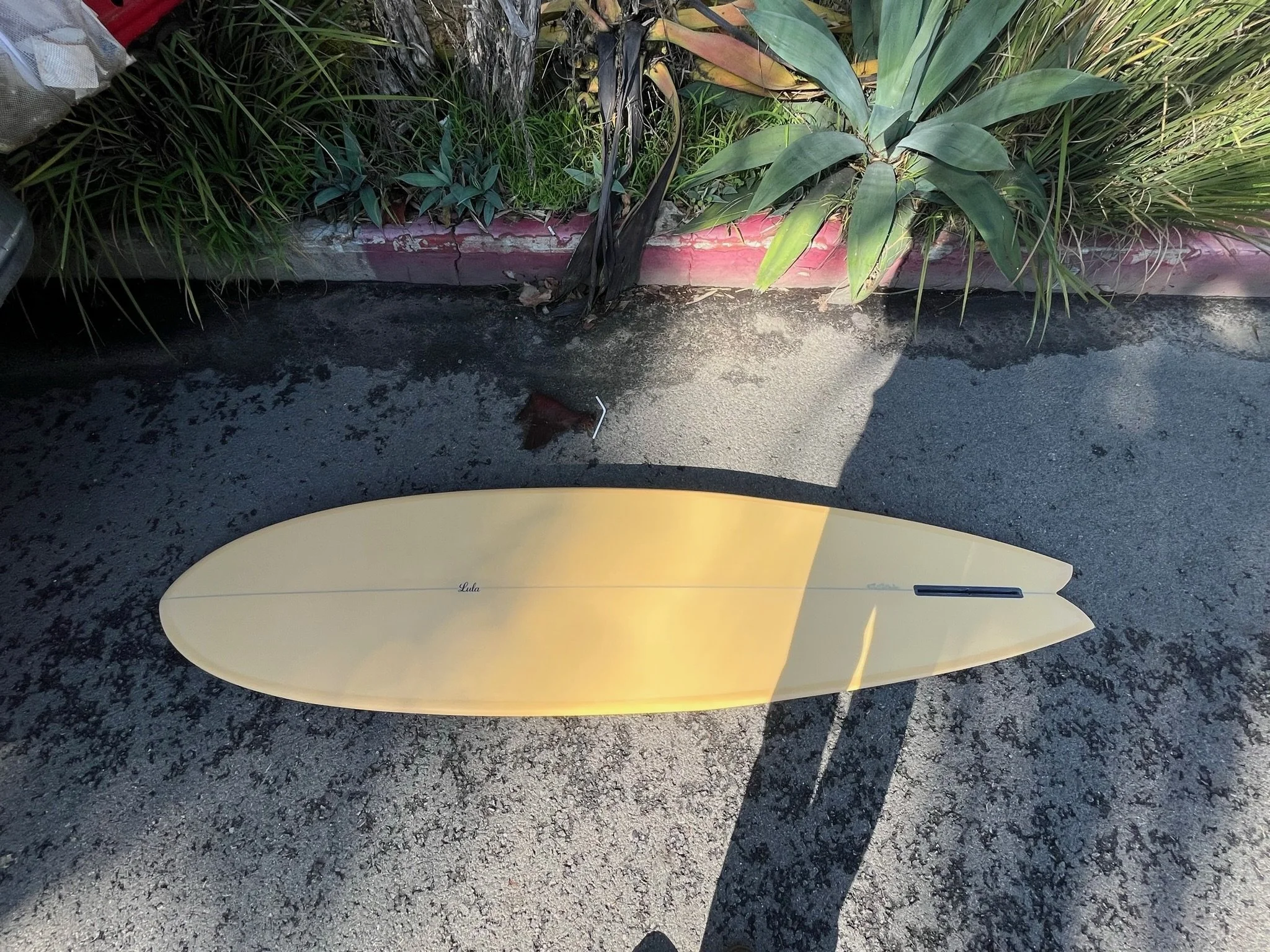 A yellow surfboard with the name Lula written on it leaning against a curb. There are plants, including long green leaves, growing over the curb and onto the sidewalk. A shadow cast by a person or object is visible on the ground.