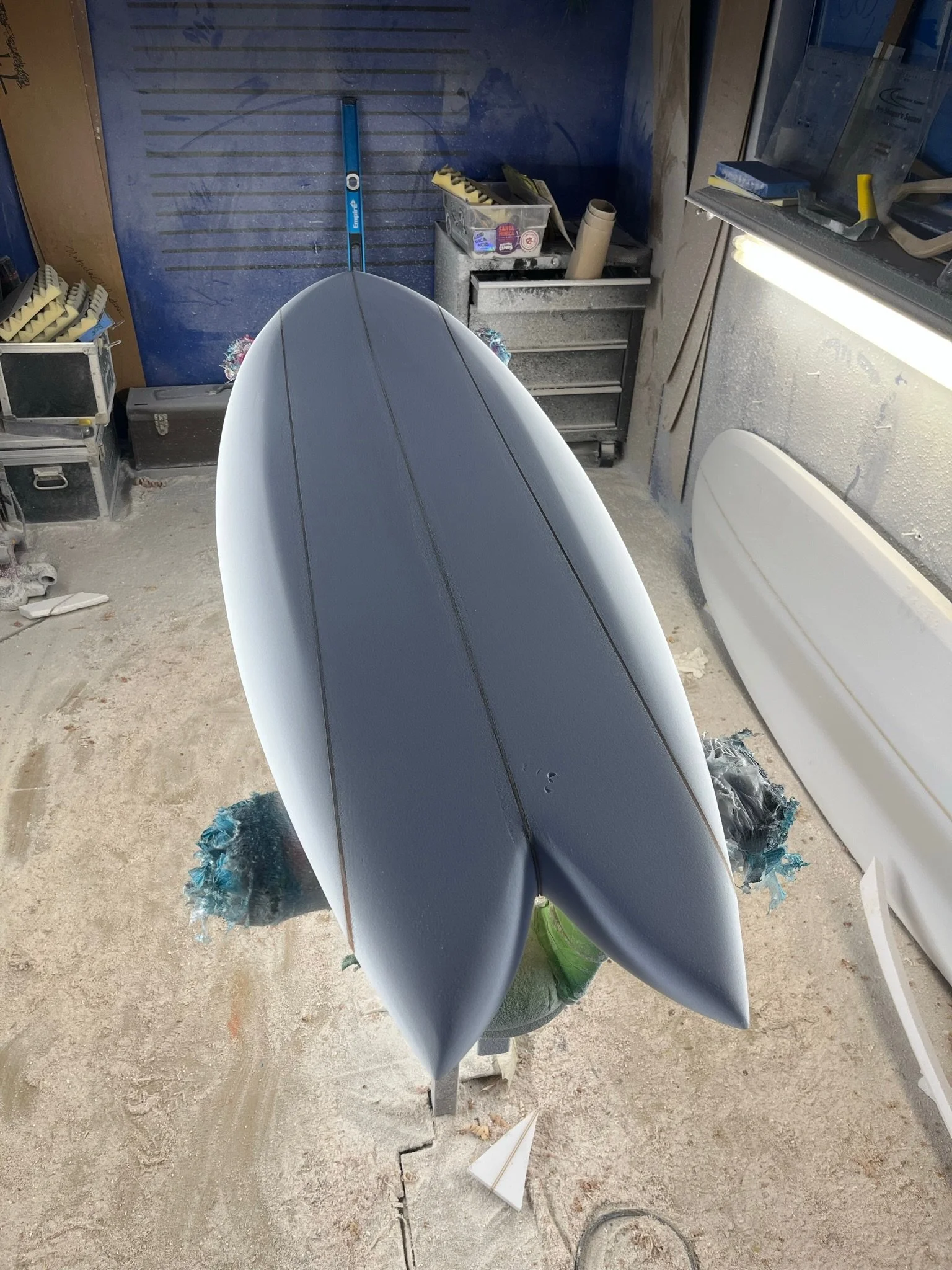 A surfboard in a workshop or garage environment, with tools and equipment in the background, leaning against a workbench, and wrapped in foam at the bottom.