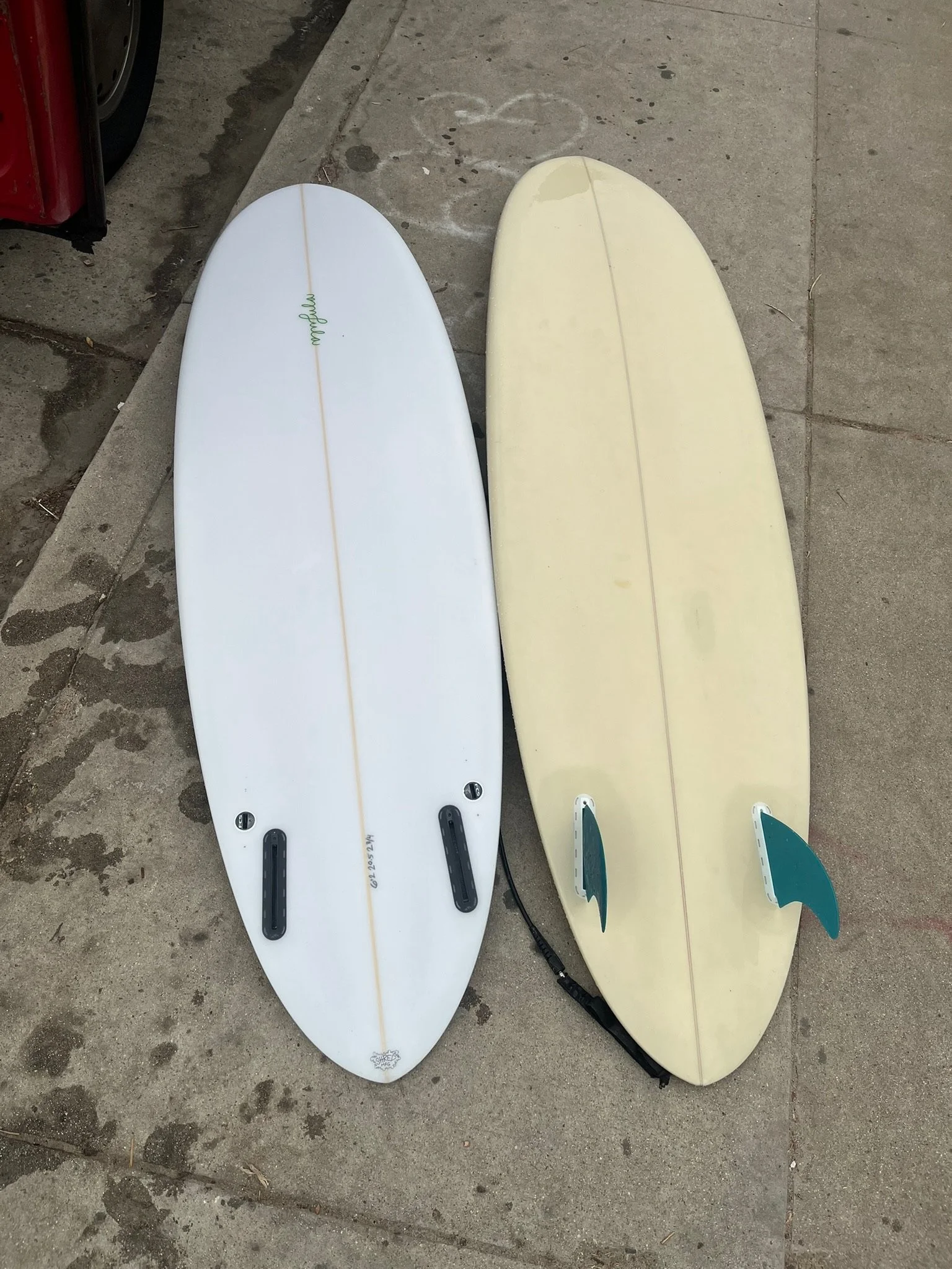 Two surfboards lying on the sidewalk, one white and one yellow, with fins attached, near a curb and a red vehicle.