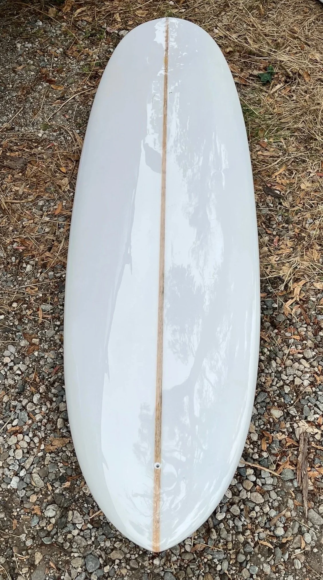 A white surfboard with a wooden stringer running down the center, placed on a gravelly patch of ground with dry leaves and twigs around.