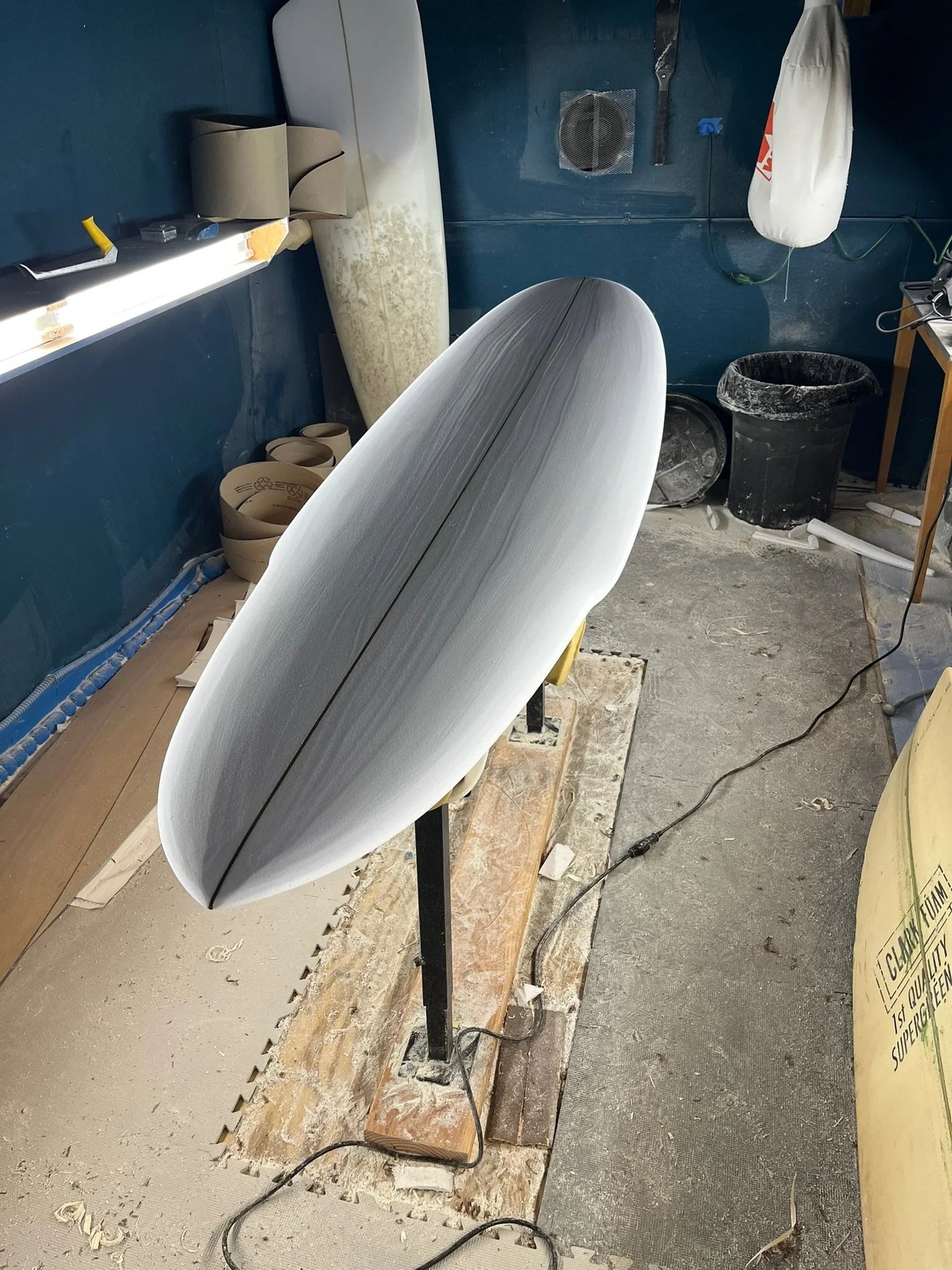 A surfboard being shaped in a workshop with sandpaper and shaping tools. The workspace is cluttered with sanding material, tools, a trash bin, and various supplies.