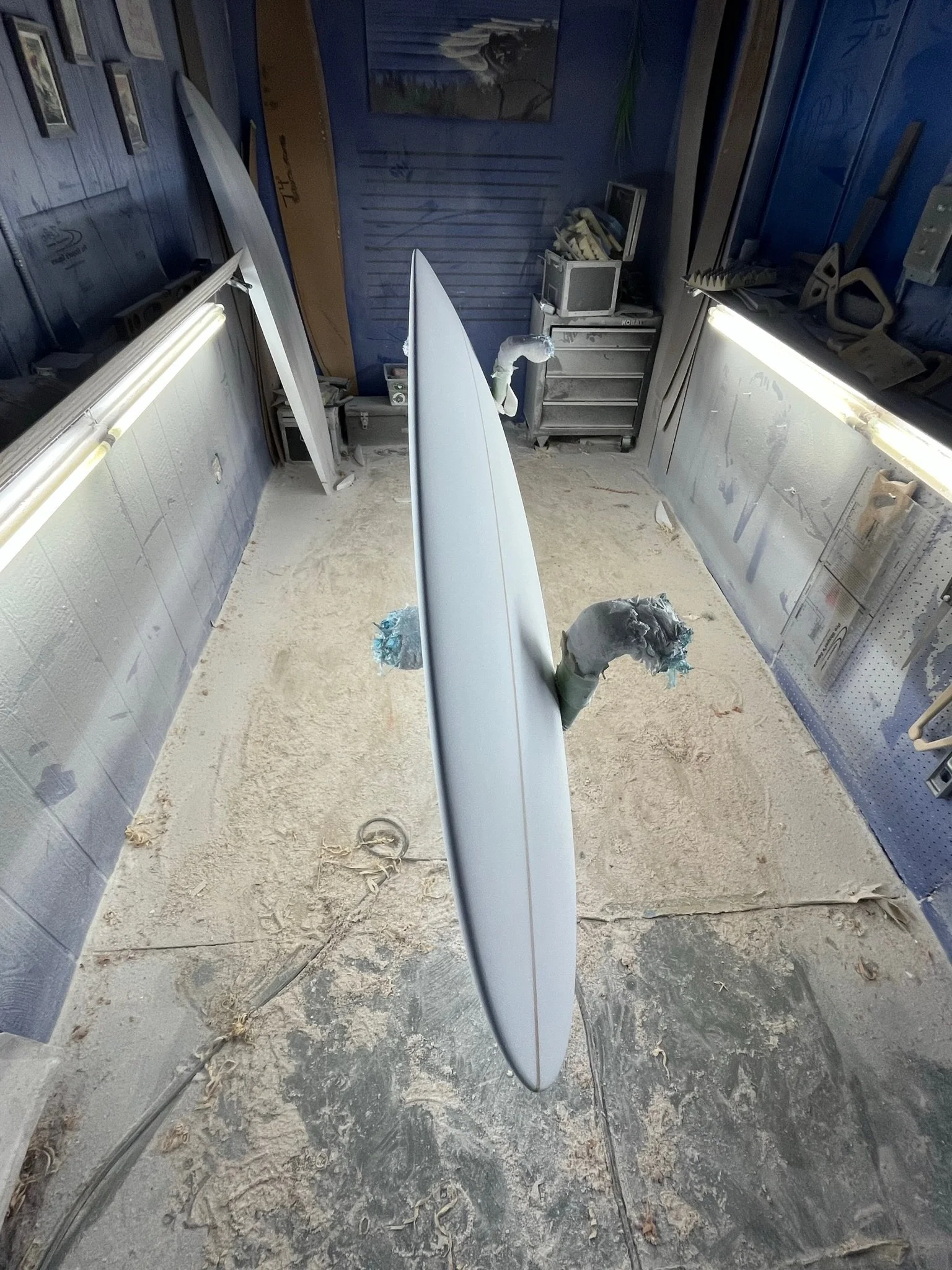 A workshop or garage with a surfboard standing upright in the center, surrounded by tools and equipment on the walls and shelves, dusty floor.