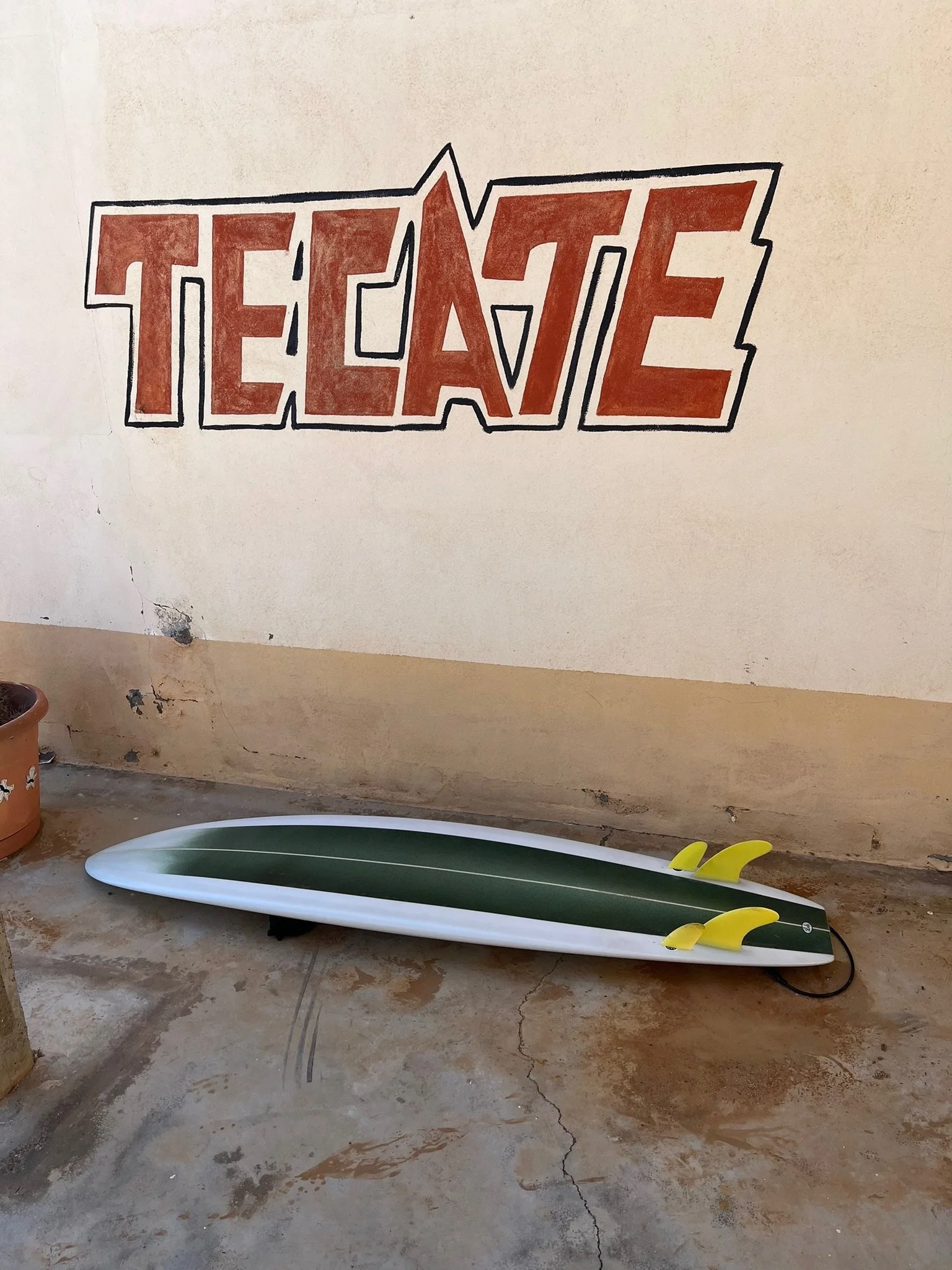 A white surfboard with yellow fins and black cord on a concrete floor beneath a wall with a colorful "TEEN C" painting.