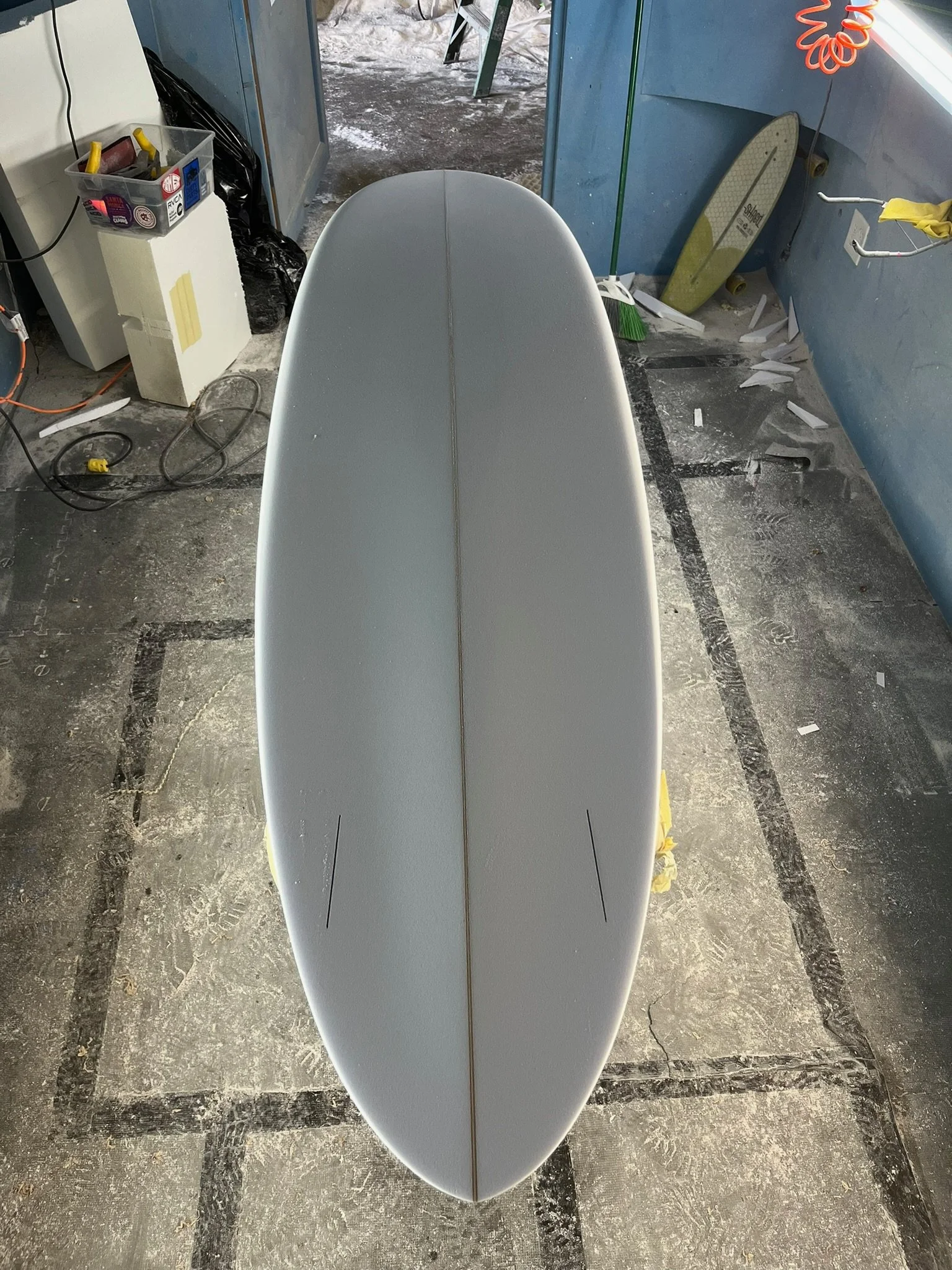 Top view of a white surfboard standing vertically on a concrete garage floor, with a blue wall and various tools and objects in the background.