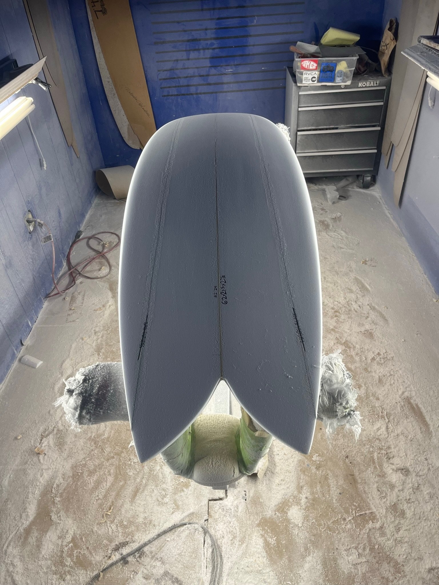 A surfboard placed upright on a padded stand in a workshop. The surfboard has a light gray top with visible seams and black markings. The workshop has a blue wall with a wavy wooden shape, a black tool chest, and various tools and supplies scattered 